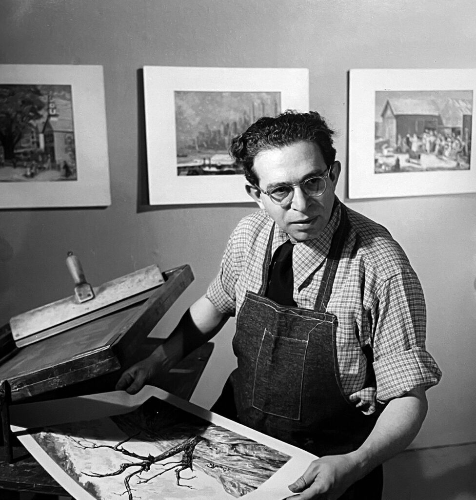 A black-and-white photo of a man in a shop apron holding a print and standing near a screen printing table