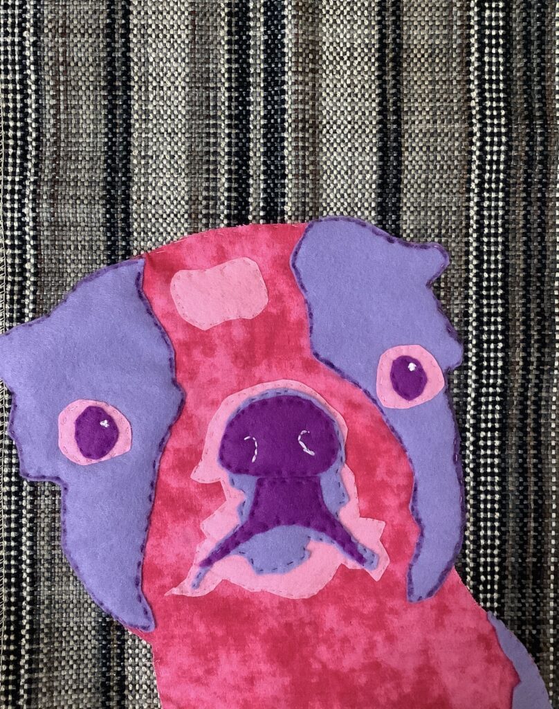 A mixed media fabric artwork depicting a pink-and-purple hued dog