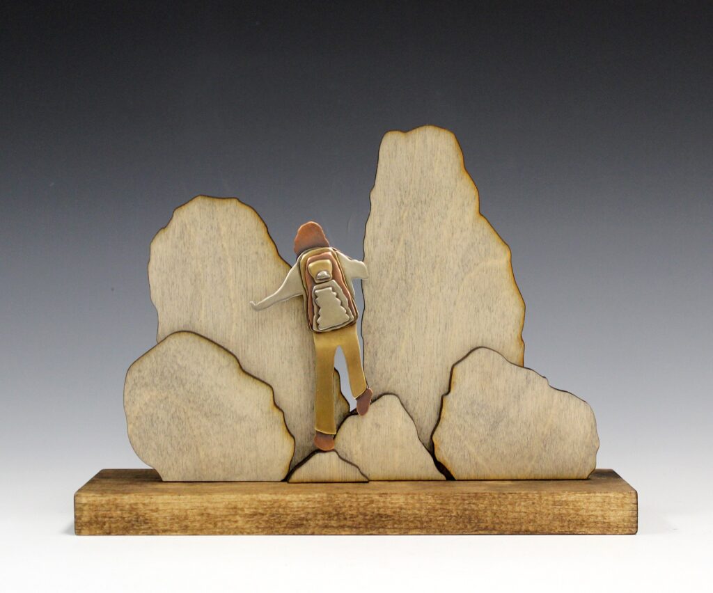 A mixed-media sculpture made of metal and wood depicting a backpacked human figure traversing boulders.