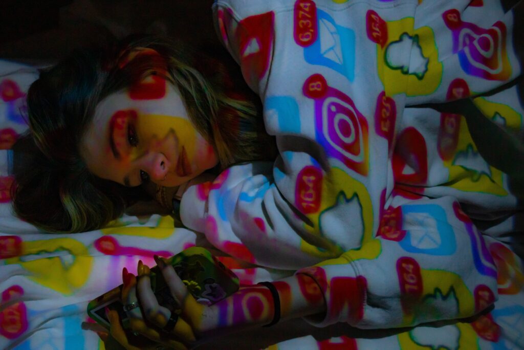 A photo of a young feminine figure laying in pajamas, using their phone; various icons of social media applications are projected over them, with notifications icons featured prominently