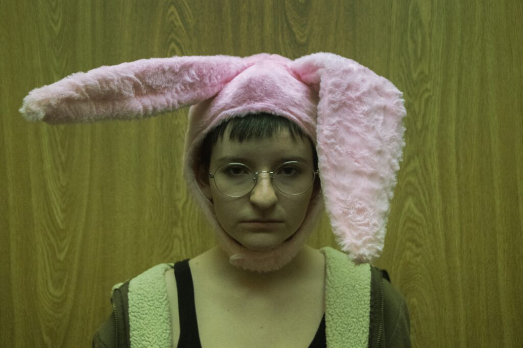 A photo of a tired-looking young person with glasses and large, punk, fluffy bunny ears, standing in front of a wooden backdrop